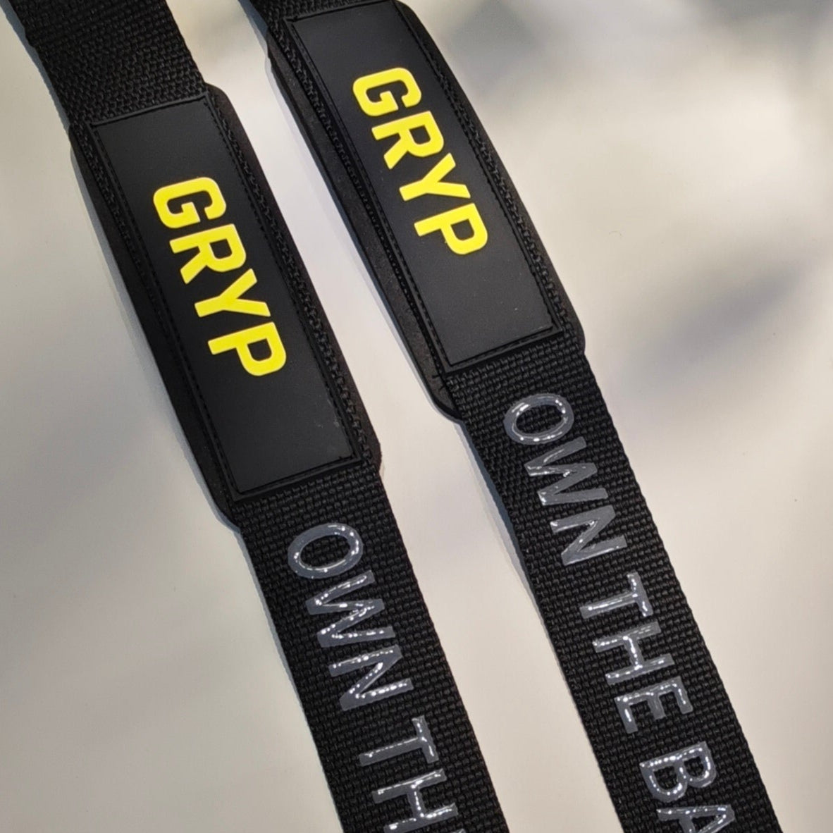 Detalle de dos straps GRYP negros con el logo amarillo y el texto OWN THE BAR en gris.